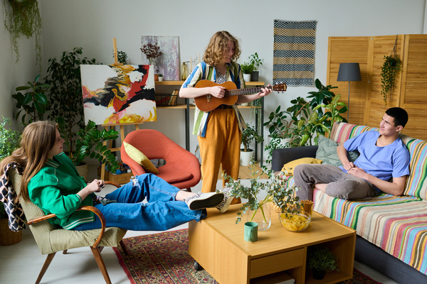 Young friends dressed in bright modern outfits sit and listen to their curly-haired girlfriend playing guitar standing in the middle of a bright room with a lot of plants
